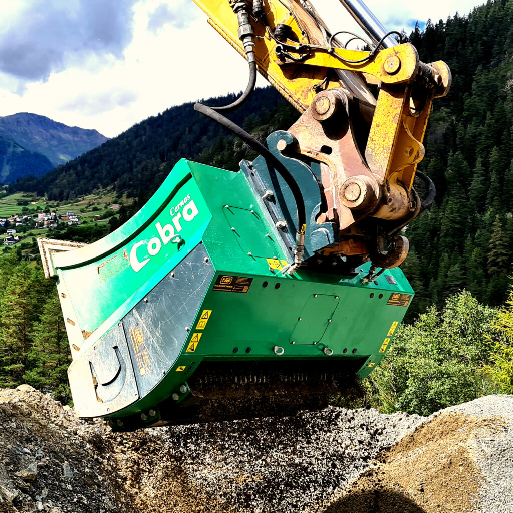 Ein gelber Baggerarm hält einen grünen Cobra-Anbau, der auf einer Schotterfläche Gestein zerkleinert, vor alpiner Bergkulisse.