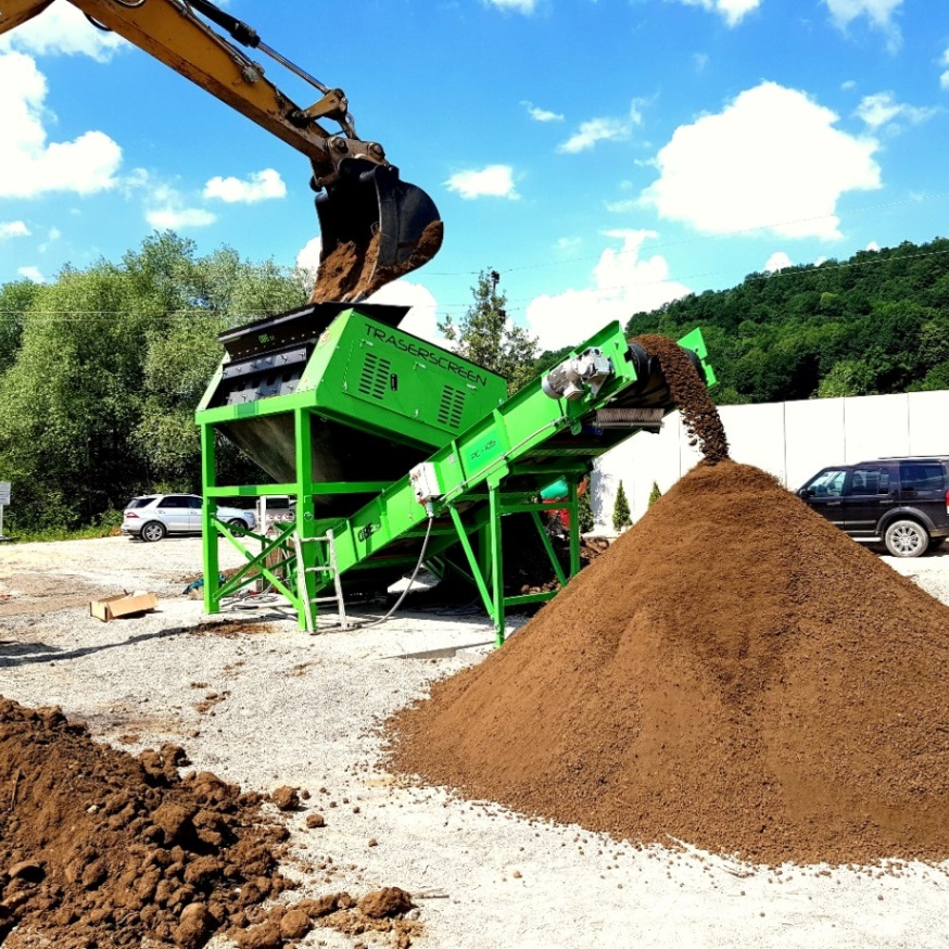 Ein grüner TRASERSCREEN-Siebmaschine fördert Erdmaterial auf einen großen Haufen, während ein Baggerlöffel von oben nachführt und Fahrzeuge im Hintergrund stehen.