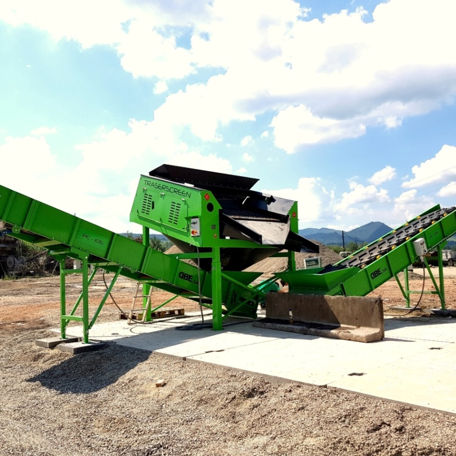 Grüne Traserscreen-Siebanlage DB-100S-2 mit schrägen Förderbändern steht auf einer Betonfläche im Freien, vor blauem Himmel und Bergen.