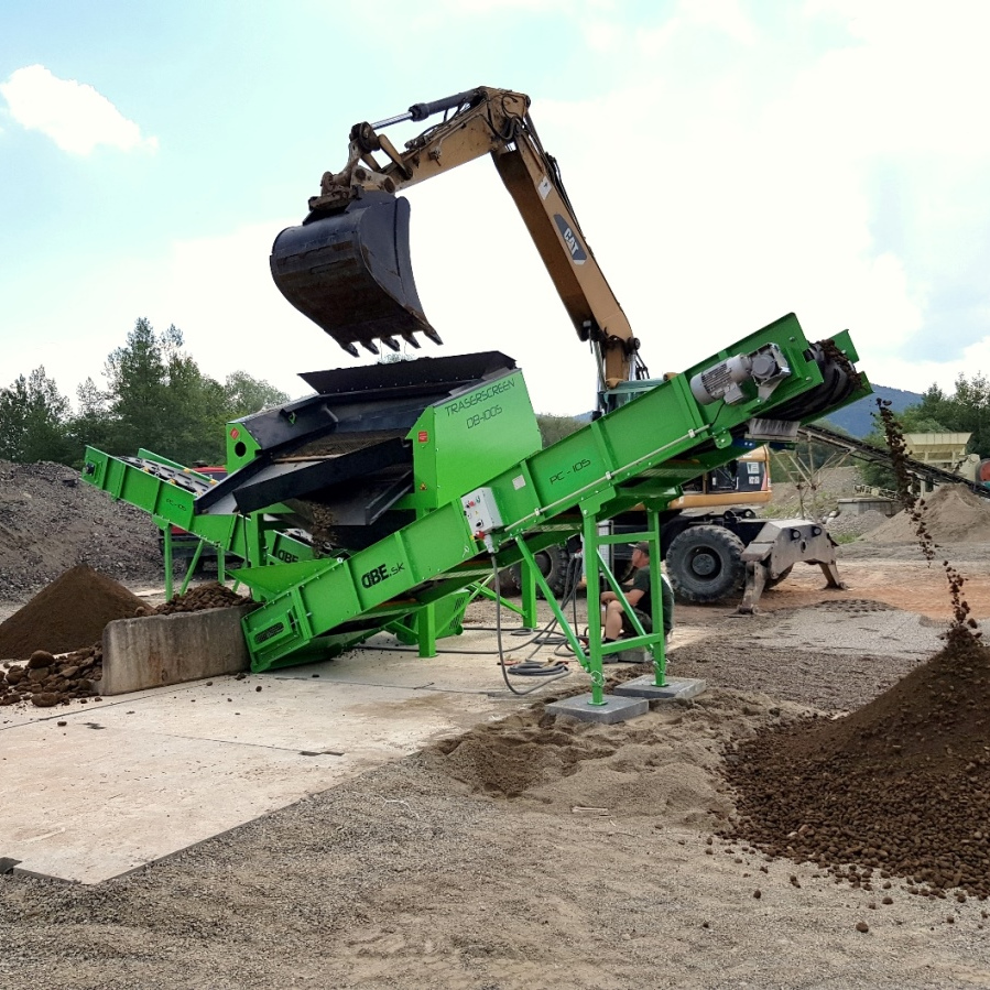 Ein gelber Bagger kippt Schutt in eine grüne TRASERSCREEN DB-100S-4 Siebanlage auf einer Baustelle, während Material auf mehrere Förderbänder läuft.