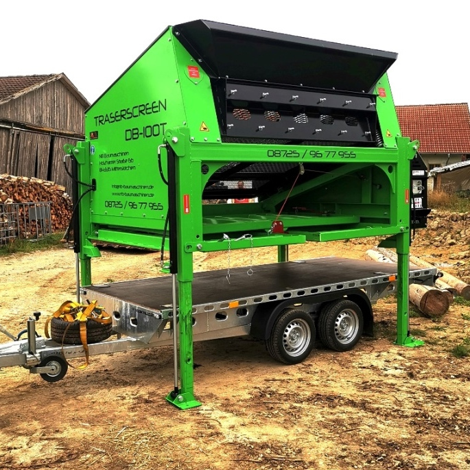 Ein grün-schwarzer TRASERSCREEN DB-100T steht auf einem Anhänger im Freien, mit sichtbaren Stützbeinen, Zaun, Holzstapeln und Erdhintergrund bei einem Hof.