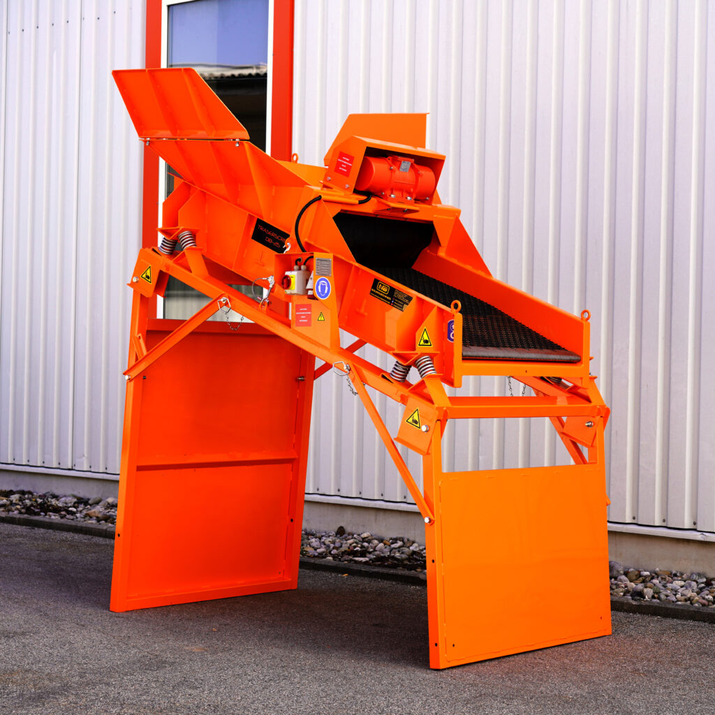 Vor einer weißen Metallwand steht eine leuchtend orange Siebmaschine mit Förderband, Klappen und Warnaufklebern im Freien für den Recyclingbetrieb.