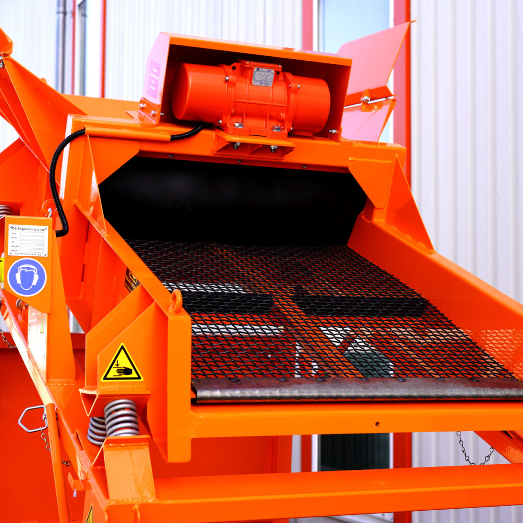 Leuchtend orangefarbener DB-25-4 mit Gitterband, Antriebsmotor und Schutzhauben vor heller Hallenwand, dazu Warnsymbol und Federn sichtbar.