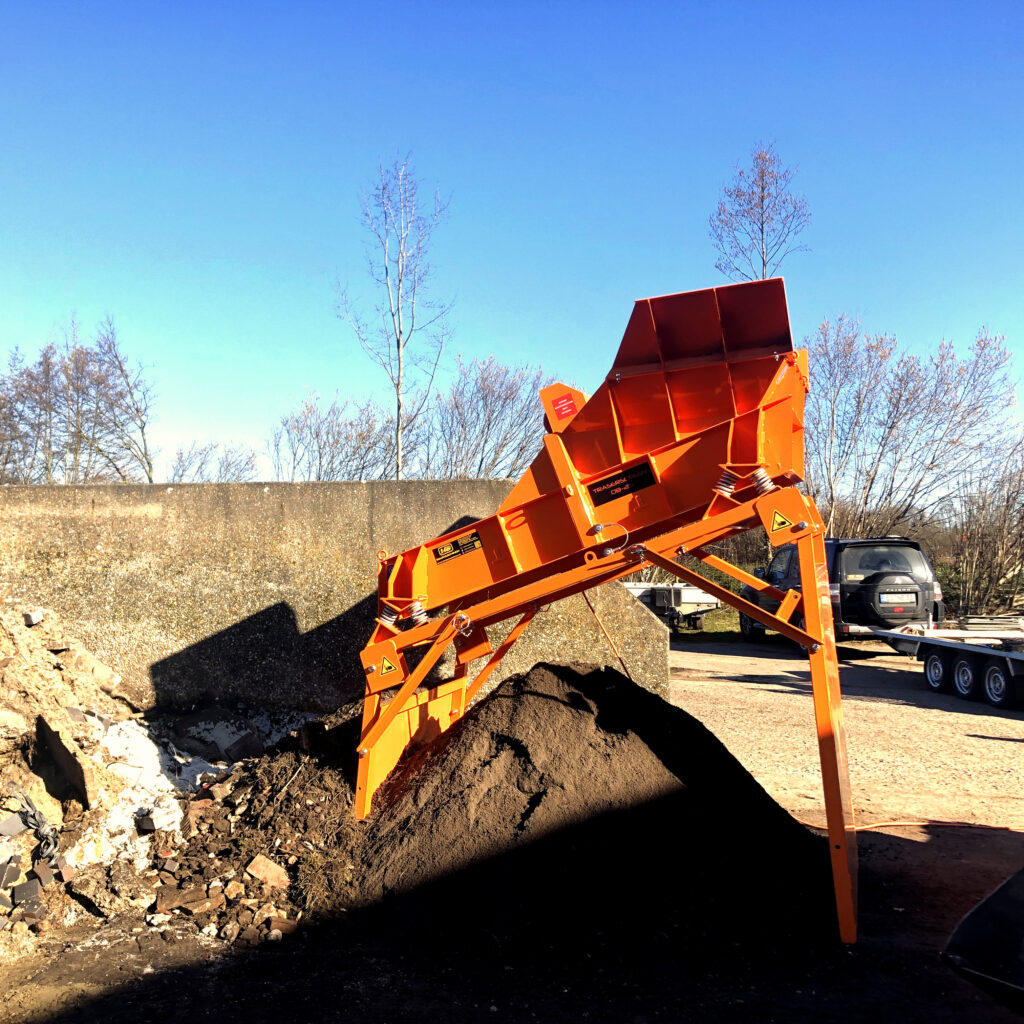 Großer orangefarbener Siebmaschinen-Aufbau kippt Material zu einem dunklen Haufen vor einer Betonwand, dahinter stehen kahle Bäume und ein SUV.
