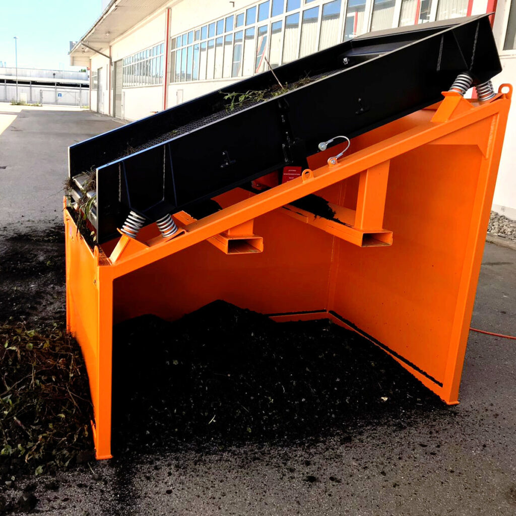 Ein orange-schwarzer Industrieschüttcontainer mit geöffneter Klappe steht auf einem Werksgelände, gefüllt mit dunklem Recyclingmaterial und Pflanzenresten.