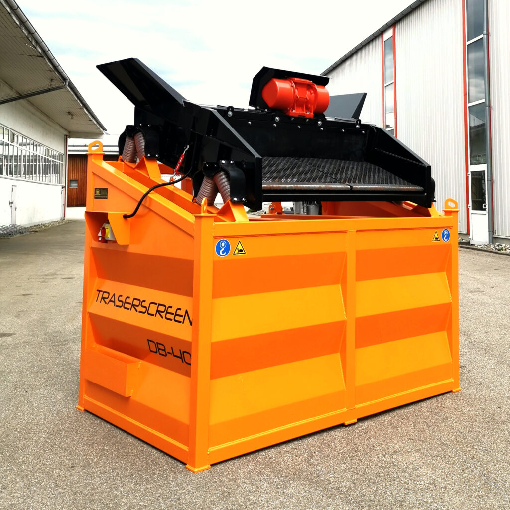 Orange-schwarze Traserscreen DB-40-7 Siebanlage steht auf einem Werksgelände zwischen Hallen, mit Fördertechnik, Gitterfläche und rotem Antrieb oben.