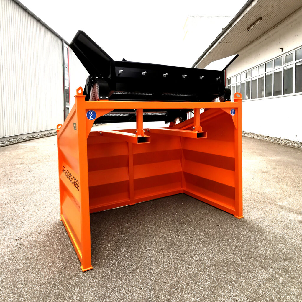 Ein orange-schwarzer Trenn- und Sortierbehälter mit geneigtem Oberteil und robustem Stahlrahmen steht zwischen zwei Hallen auf dem Werksgelände.