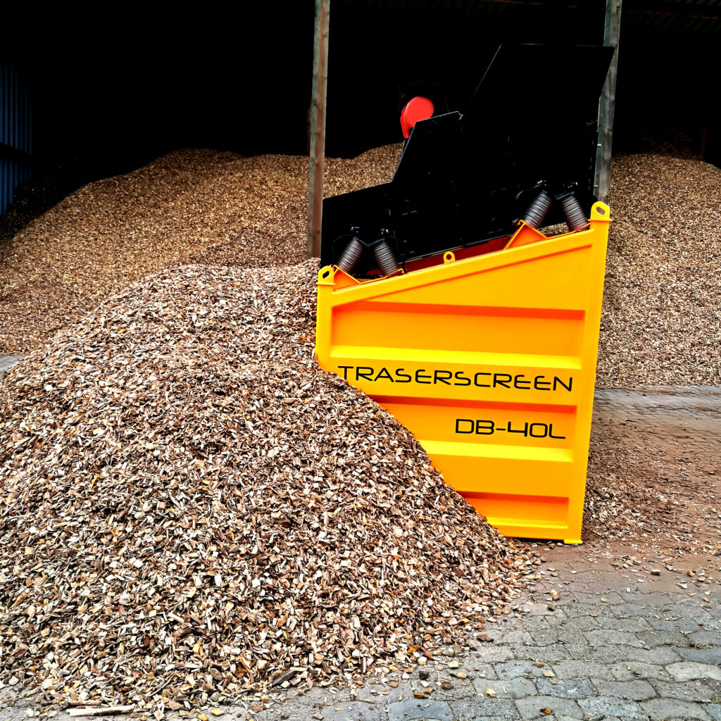 Vor einem hohen Schredderberg steht ein gelber Traserscreen DB-40L mit schwarzem Aufbau und großem Füllraum in einer dunklen Halle aus Holzschnitzeln.