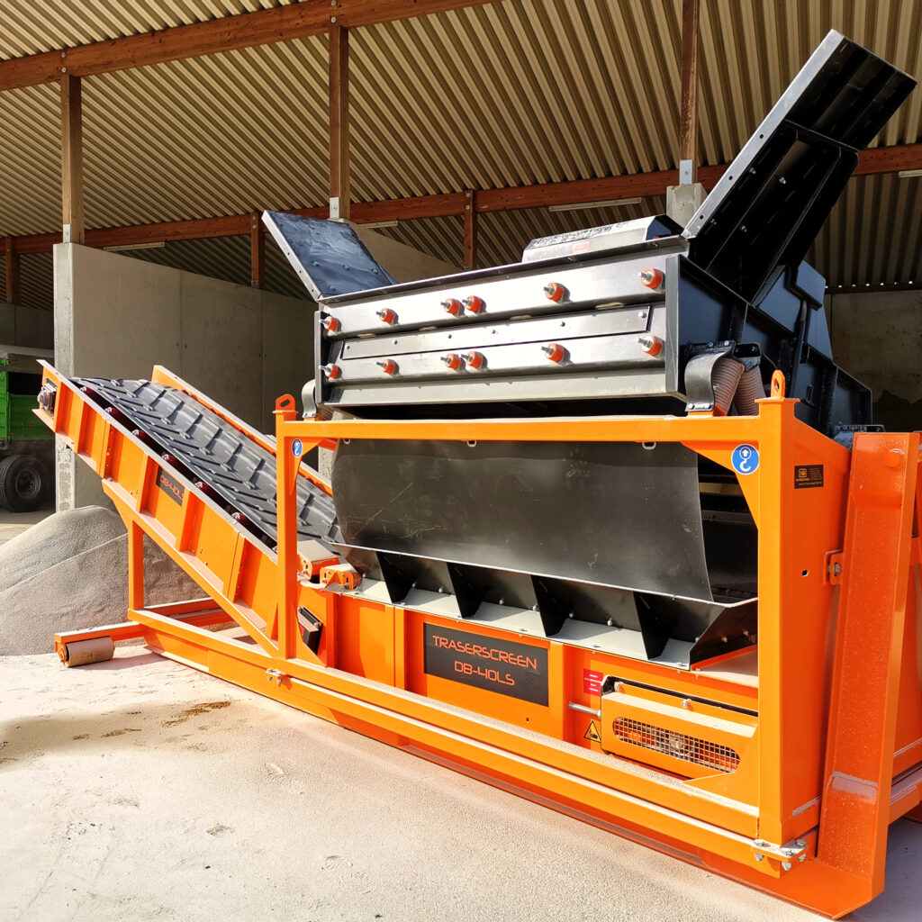 Ein orange-schwarzer Traserscreen DB-40LS steht in einer Halle, mit geneigtem Förderband, großem Siebkasten und sichtbaren Stützträgern im Licht.