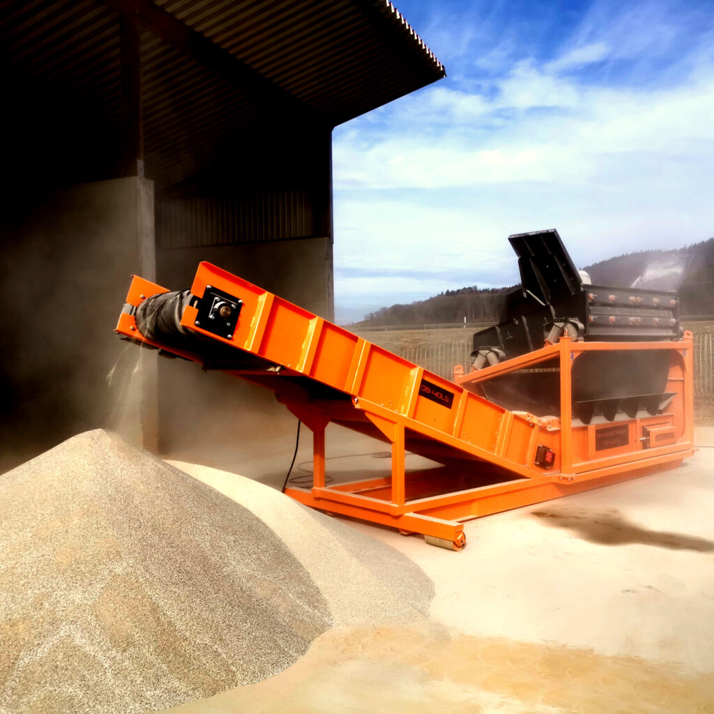 Eine orange Förderbandanlage mit schwarzer Siebeinheit verteilt Schüttgut neben einem großen Haufen unter freiem Himmel vor hügeliger Landschaft.