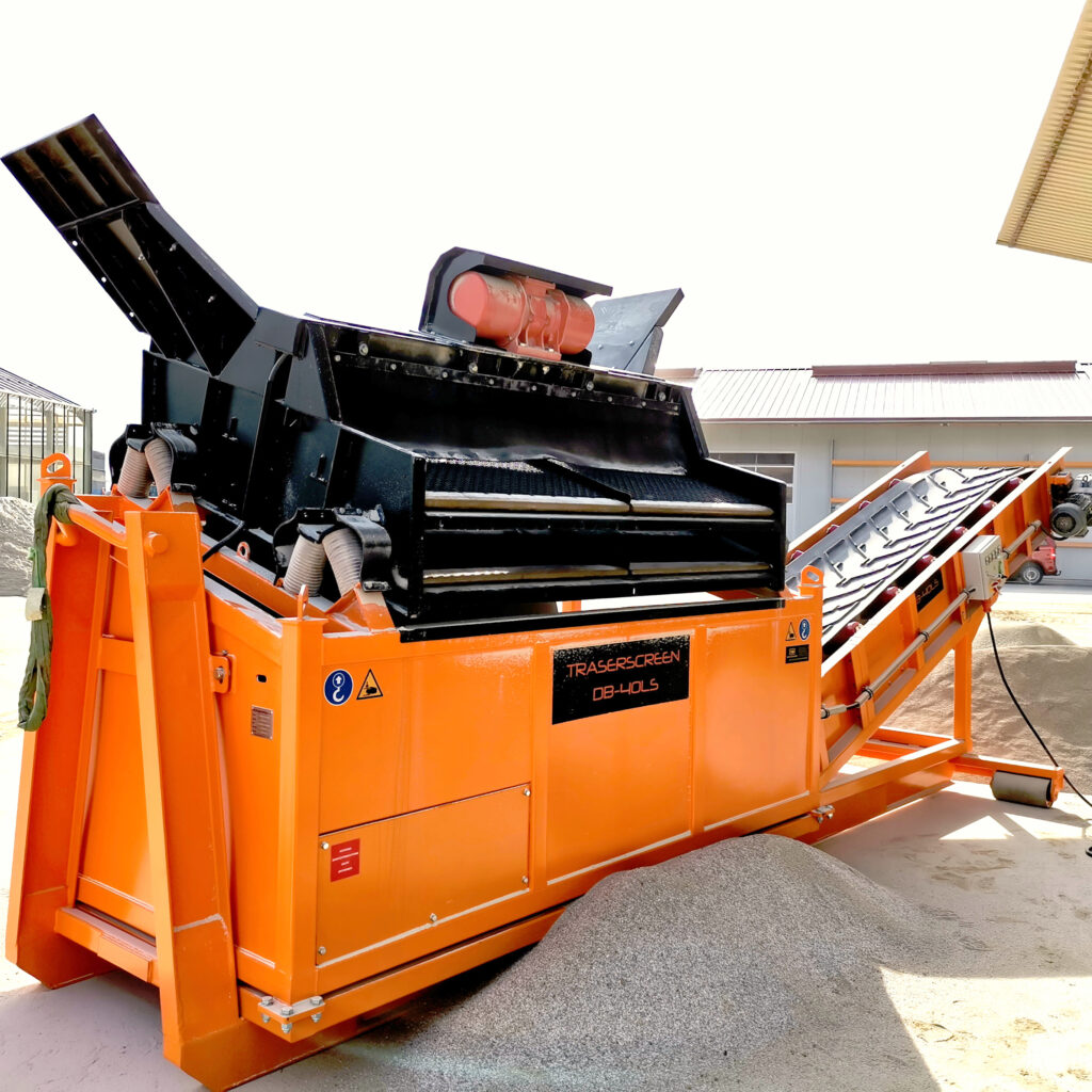 Eine orange-schwarze Traserscreen DB-40LS steht im Hof vor einer Halle, mit Förderbändern und Siebdecks, während helles Material darunter liegt.
