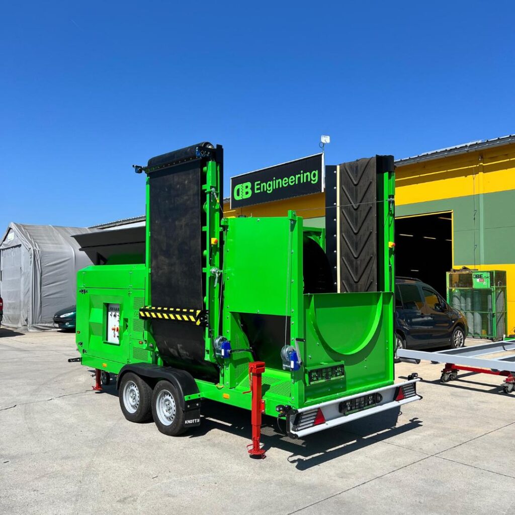 Ein grünes Recyclingmaschinen-Anhängerfahrzeug mit schwarzem Förderband und Warnstreifen steht auf dem Hof vor einer Werkhalle unter klarem Himmel.