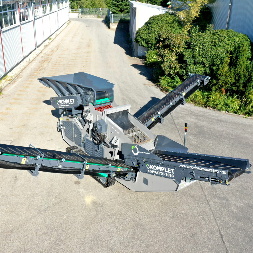 Ein grauer Komplet Kompatto 5030 zerkleinert Material auf einem sonnigen Betriebshof, während Förderbänder und Trichter klar zu sehen sind.