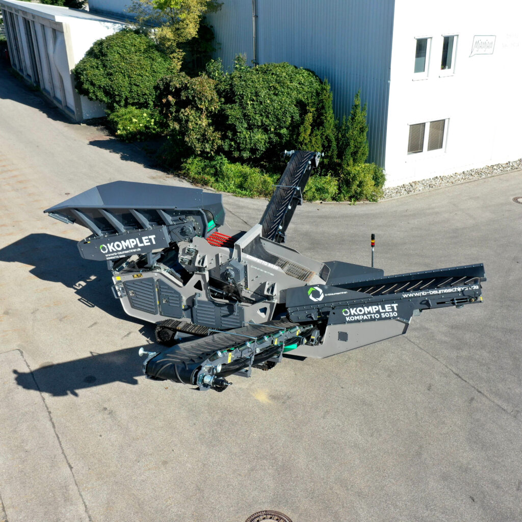 Auf einem Asphaltgelände steht ein grauer und schwarzer Komplet Kompattto 5030 mit Förderband und Trichter neben einem Industriegebäude in der Sonne.