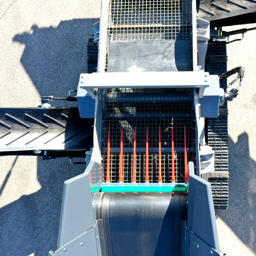 Blick von oben auf eine große Recyclingmaschine mit Gitterrosten, Fördertechnik und Förderbändern in Grau und Schwarz, umgeben von hellem Asphalt.