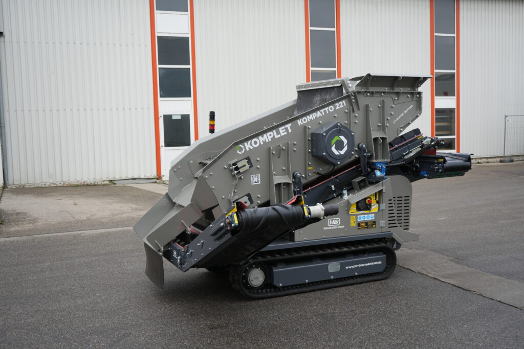 Ein grauer Komplet KOMPATTO 221 Brecher mit Förderband und Raupenfahrwerk steht vor einer weißen Industriehalle mit orange umrahmten Fenstern.