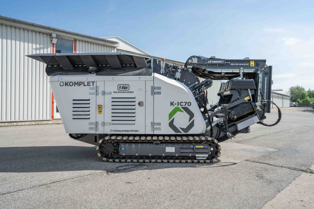 Auf dem Werksgelände steht ein grauer Komplet K-IC70 mit schwarzem Förderarm, Kettenfahrwerk und offenen Wartungsklappen vor einer Halle im Sonnenschein.