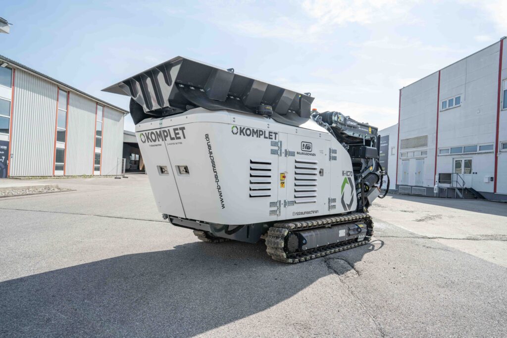 Ein weiß-schwarzer KOMPLET K-JC70 auf Raupen steht zwischen Industriegebäuden auf dem Hof von NB Baumaschinen unter hellem Himmel.