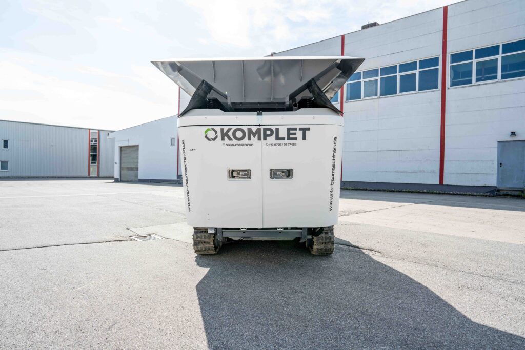 Großer weißer Komplet-Recyclingmaschinen mit Fronttrichter und Kettenfahrwerk steht vor einer hellen Industriehalle auf sonnigem Asphalt.