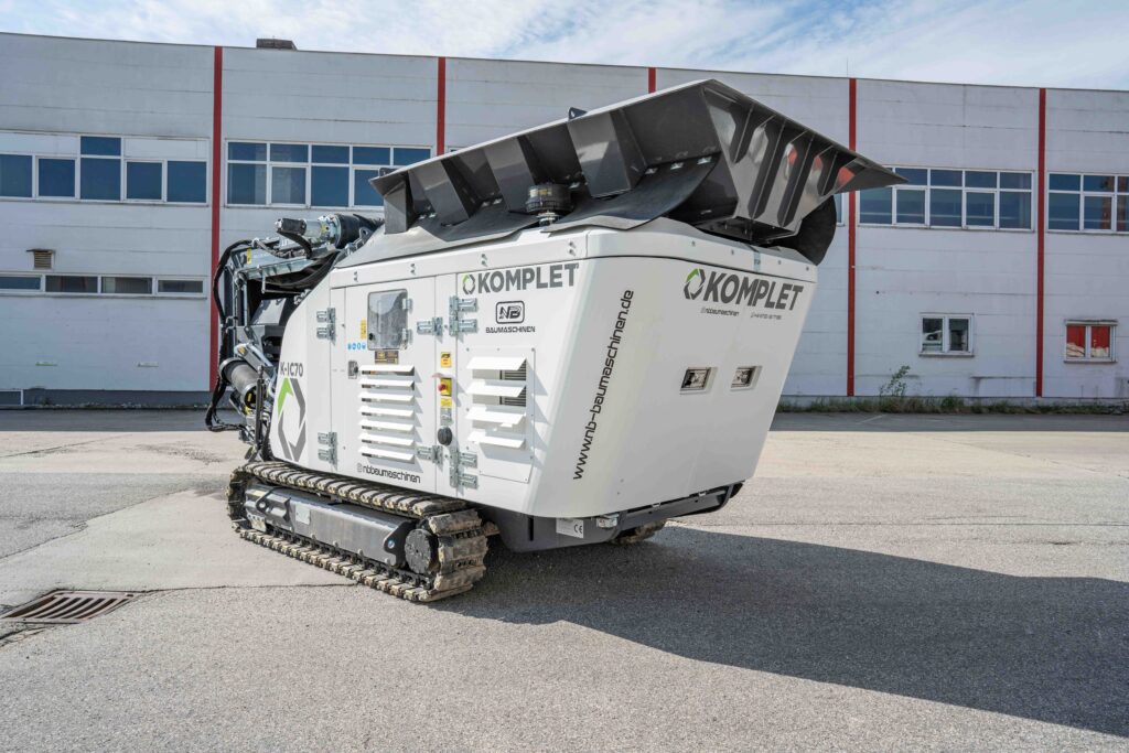 Ein weißer KOMPLET-Recyclingbrecher mit schwarzem Aufgabetrichter steht auf Raupen vor einer Industriehalle und zeigt seitlich NB-Baumaschinen-Beschriftungen.