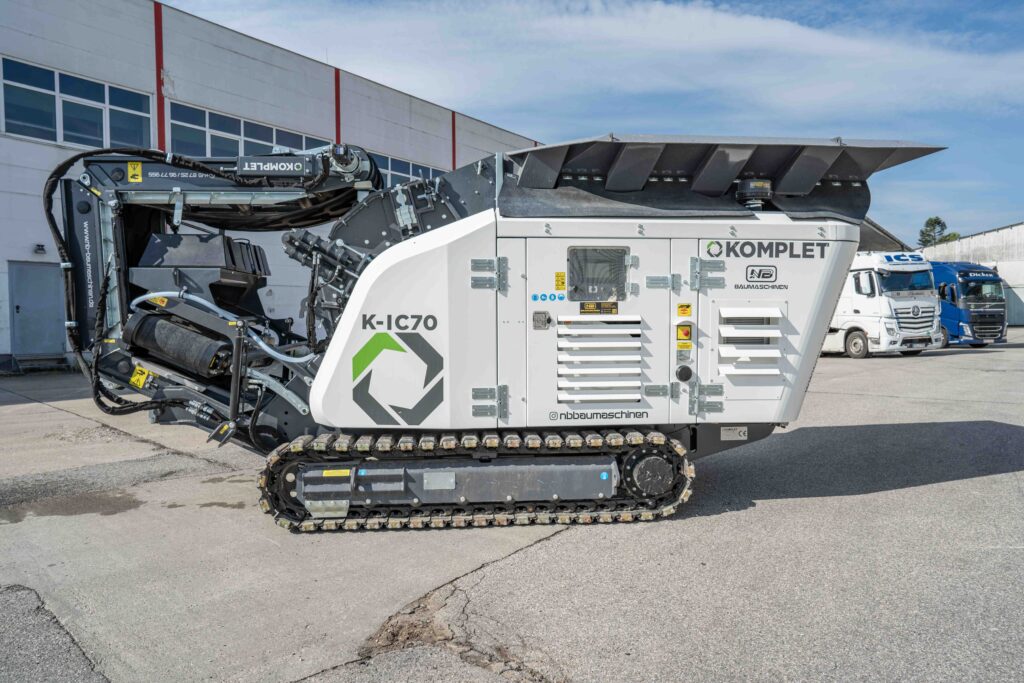 Ein weiß-schwarzer Kompakt-Recycler K-IC70 von KOMPLET steht auf Ketten vor einem Betriebsgebäude, während im Hintergrund Lkw parken.