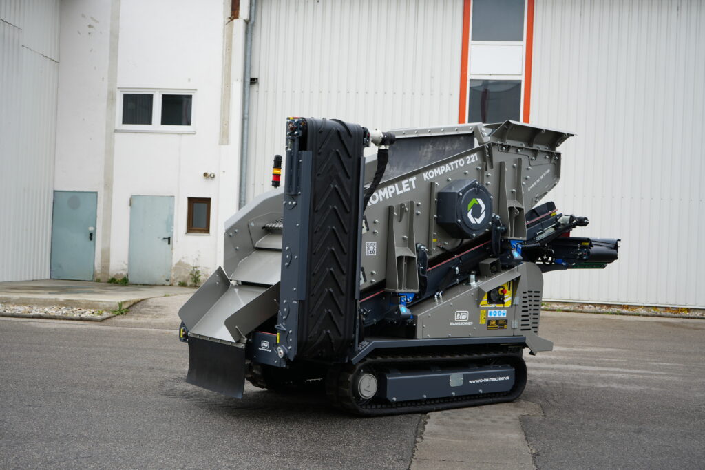 Vor einer hellen Industriehalle steht ein kompakter, grauer Komplet Kompatto 221 mit Förderband, schwarzem Transportband und Kettenfahrwerk.
