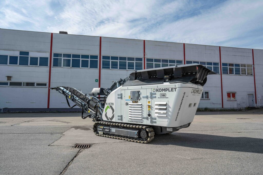 Ein weißer KOMPLET K-IC70 Recyclingbrecher mit schwarzem Austragsband steht vor einer hellen Industriehalle auf dem Asphalt im Freien.