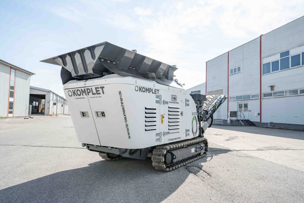 Ein weißer Kettenbrecher von Komplet steht auf einem Industriehof zwischen Hallen, mit schwarzem Einfülltrichter und Förderband unter hellem Himmel.