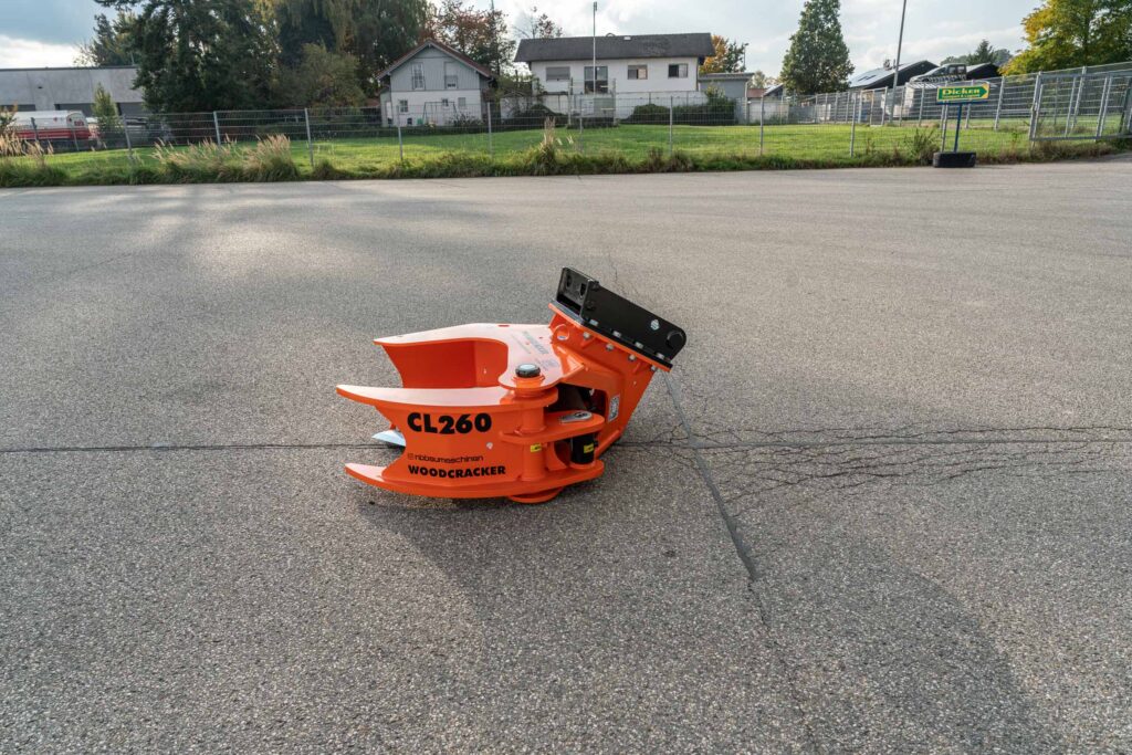 Ein orangefarbener WOODCRACKER CL260 mit schwarzer Aufnahme steht auf grauem Asphalt vor einem eingezäunten Gelände in ruhiger Tageslichtszene.