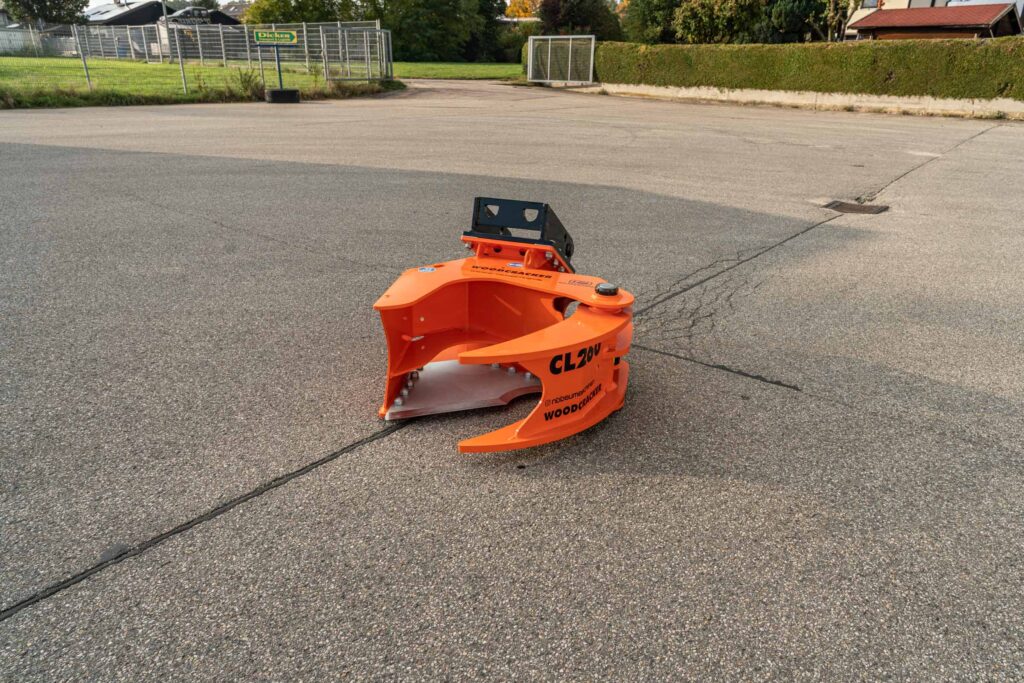 Orangefarbener WOODCRACKER CL20V auf Asphalt, mit schwarzer Aufnahme oben und breiter Greiföffnung, vor einem eingezäunten Gelände im Freien.