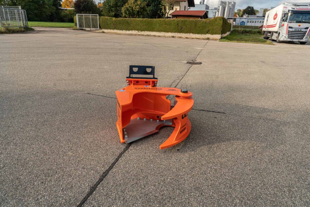 Ein orangefarbener Woodcracker-Anbau sitzt auf dem Asphalt, im Hintergrund stehen ein DAF-Lkw, Hecken und Industriegebäude auf dem Hof.
