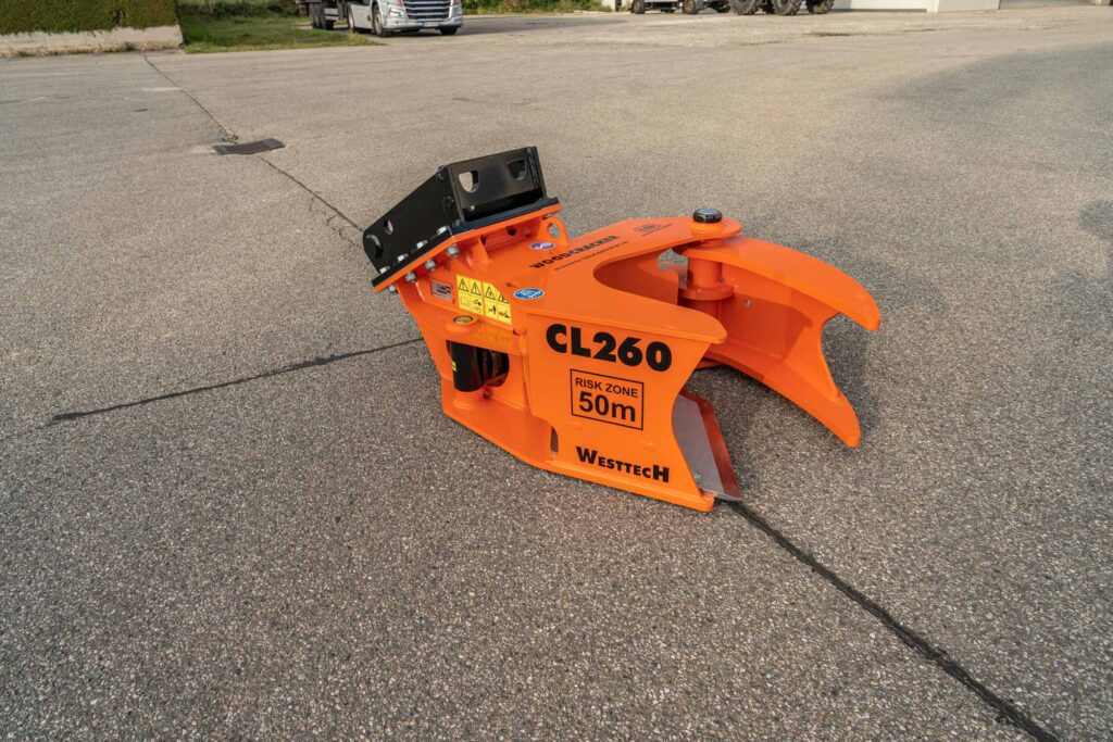Ein orangefarbener Westtech CL260 Holzgreifer liegt auf einem Asphaltplatz, mit schwarzer Aufnahme, Warnhinweis 50 m und sichtbarer Markenbeschriftung.