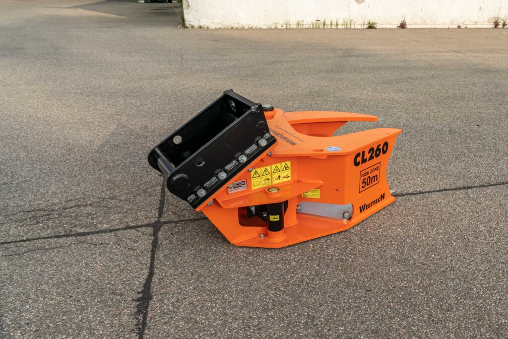 Ein orange-schwarzer Westtech CL260 Anbaugreifer liegt auf Asphalt, mit sichtbaren Warnaufklebern und großem schwarzen Aufnahmebügel.