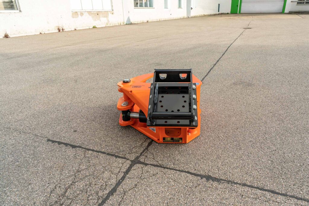 Ein orange-schwarzer Hydraulikgreifer für Baumaschinen steht auf rauem Asphalt vor einer weißen Hallenwand mit grünen Türen und Fenstern.