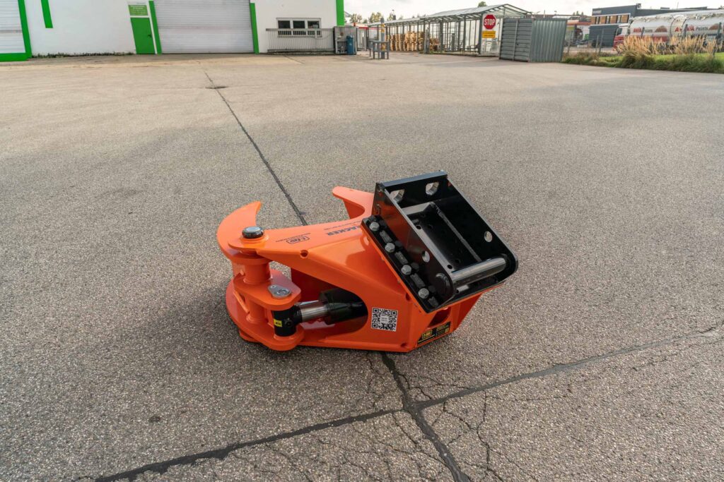Ein orange-schwarzer Anbaugreifer mit Hydraulikzylinder liegt auf einem Asphaltplatz vor einer Industriehalle und einem Stoppschild.