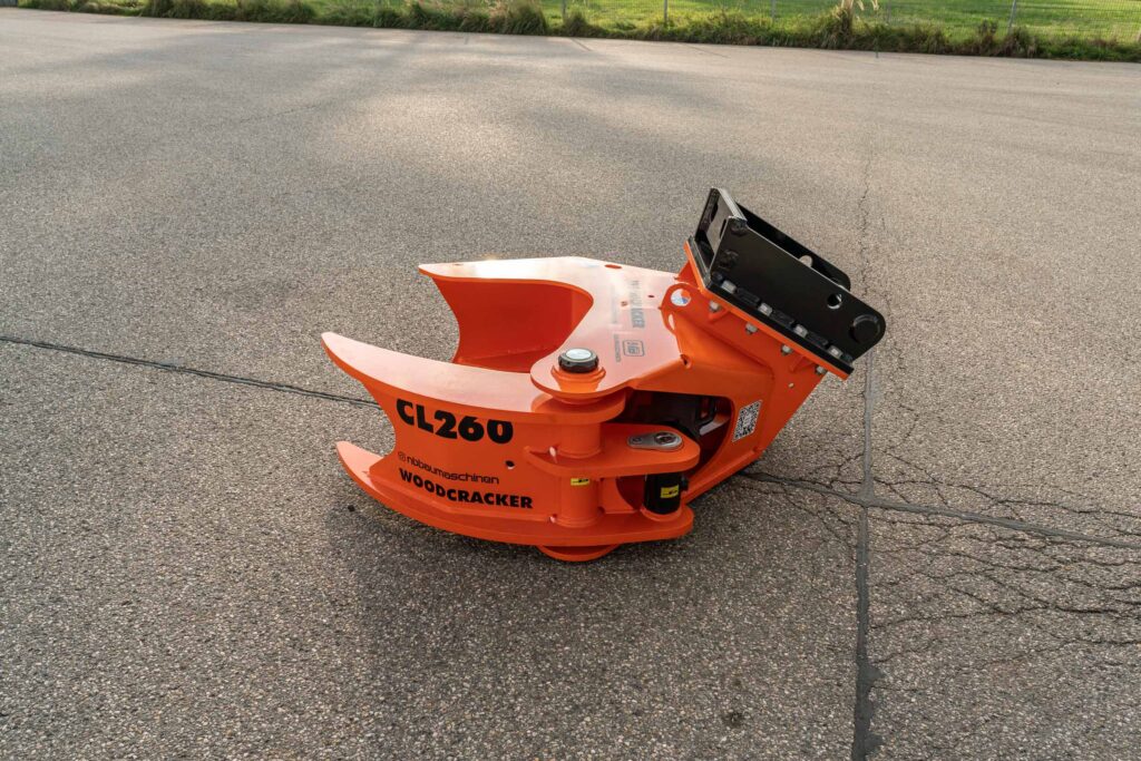 Orangefarbener Woodcracker CL260 mit schwarzer Aufnahme liegt auf rauem Asphalt; die massive Schneid- und Greifkonstruktion ist klar sichtbar, im Hintergrund Wiese und Zaun.