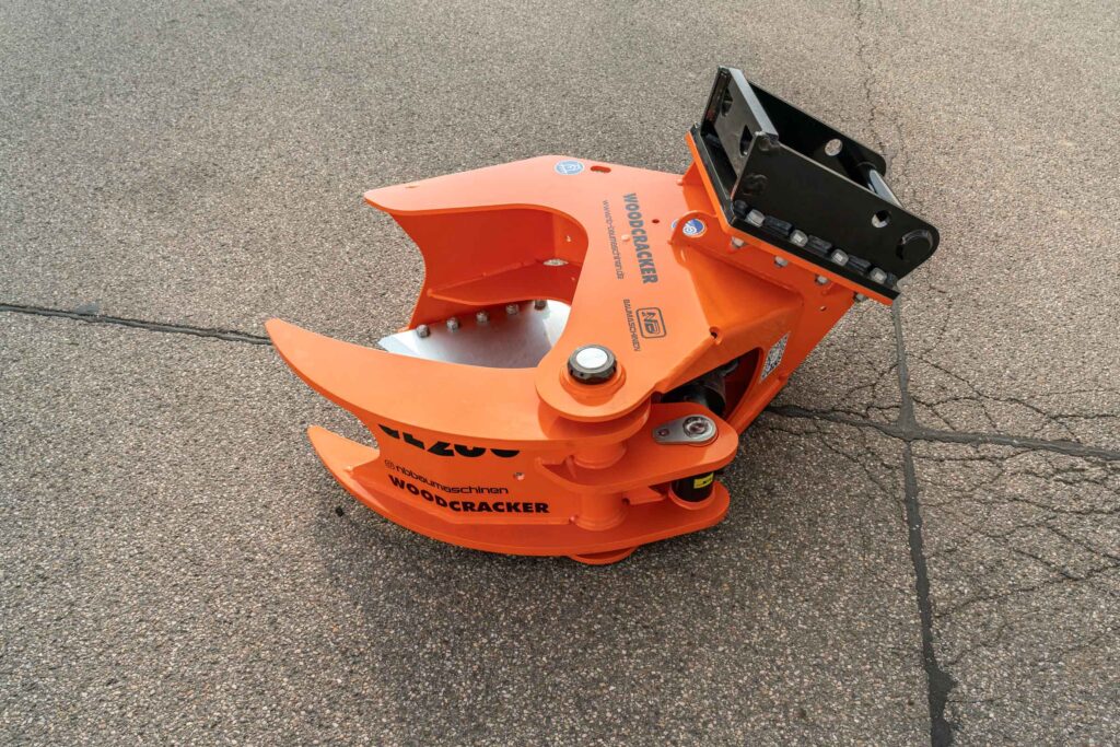 Ein orange-schwarzer WOODCRACKER-Anbaugreifer liegt auf Asphalt; der massive Stahlkörper zeigt die Marke NB Baumaschinen und die robuste Bauweise.