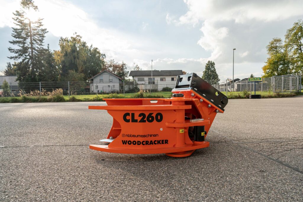 Orange-schwarzer WOODCRACKER CL260 mit massiven Stahlarmen steht auf einem Asphaltplatz vor Zaun, Bäumen und Wohnhäusern unter hellem Himmel.