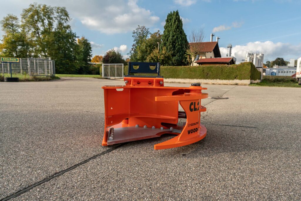 Ein orangefarbener Holzgreifer mit schwarzer Aufnahme steht auf einem Asphaltplatz, im Hintergrund sind Bäume, Gebäude und ein blauer Himmel zu sehen.