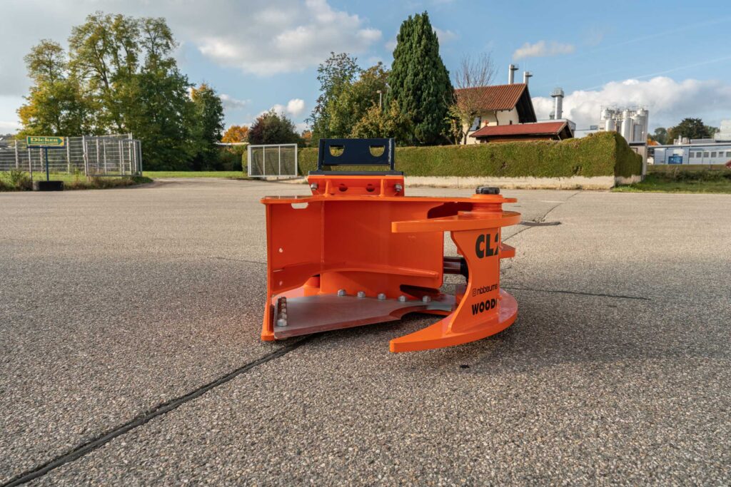 Ein orangefarbener Baumaschinen-Anbau mit schwarzer Halterung steht auf einem Asphaltplatz vor Hecken, Bäumen und einem Gebäude im Freien.