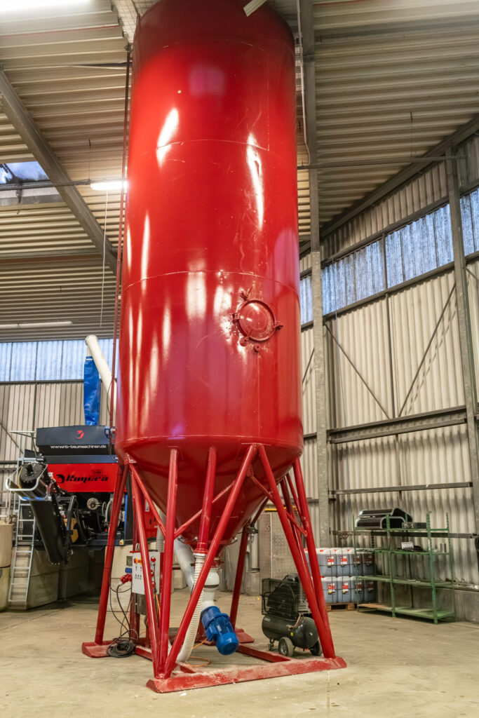 Ein hoher roter Stahltank steht in einer Industriehalle, daneben sind Metallstreben, Leitungen und Arbeitsgeräte unter dem Hallendach zu sehen.