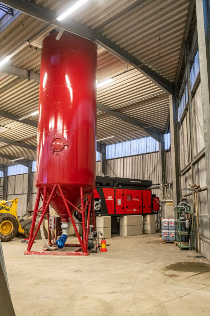 Ein hoher roter Stahltank steht in einer Werkhalle neben einer roten NB-Baumaschinen-Anlage, umgeben von Stahlträgern, Betonblöcken und Technik.