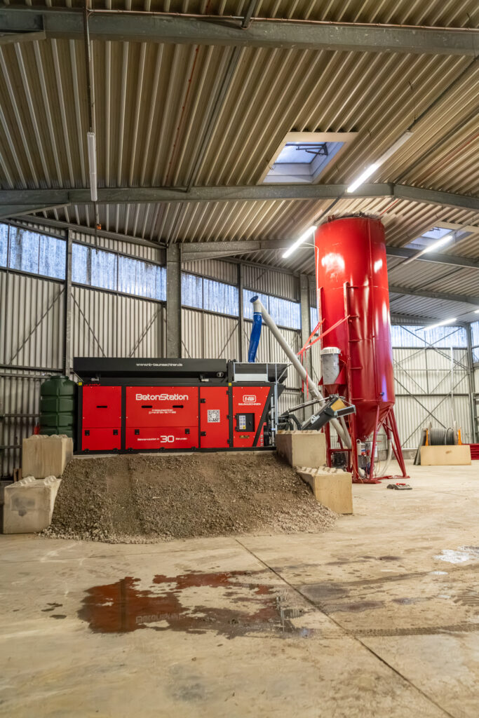 Eine rote BetonStation-Anlage von NB Baumaschinen steht in einer Industriehalle, daneben ein hoher roter Silo und ein Haufen Schotter auf dem Betonboden.