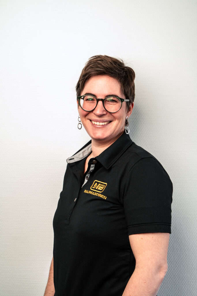 Lächelnde Mitarbeiterin mit kurzen dunklen Haaren, Brille und Ohrschmuck vor hellem Hintergrund, im schwarzen NB-Baumaschinen-Poloshirt mit gelbem Logo.