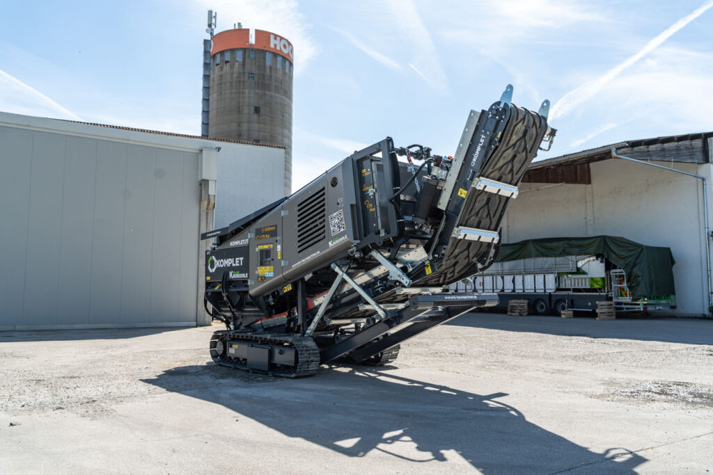 Schwarzer KOMPLET Krokodile Schredder mit Raupenfahrwerk steht auf einem Hof vor Hallen und einem Siloturm unter klarem Himmel.