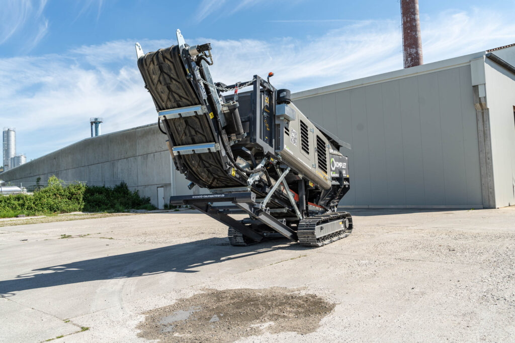Ein grauer Komplet Krokodile PLUS auf Raupenfahrwerk steht vor einer Industriehalle, mit hochgestelltem Förderband und sichtbaren Markenlogos.