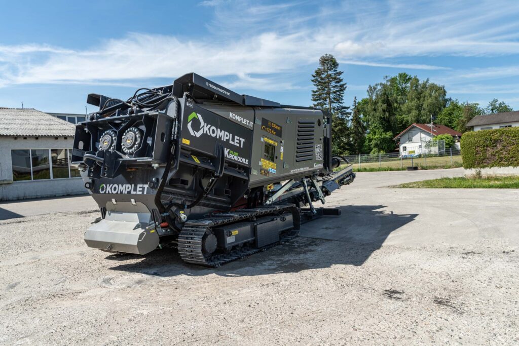 Der schwarze KOMPLET Krokodile PLUS auf Raupenfahrwerk steht auf einer Betonfläche, mit sichtbaren Hydraulikleitungen, grobem Chassis und blauem Himmel.