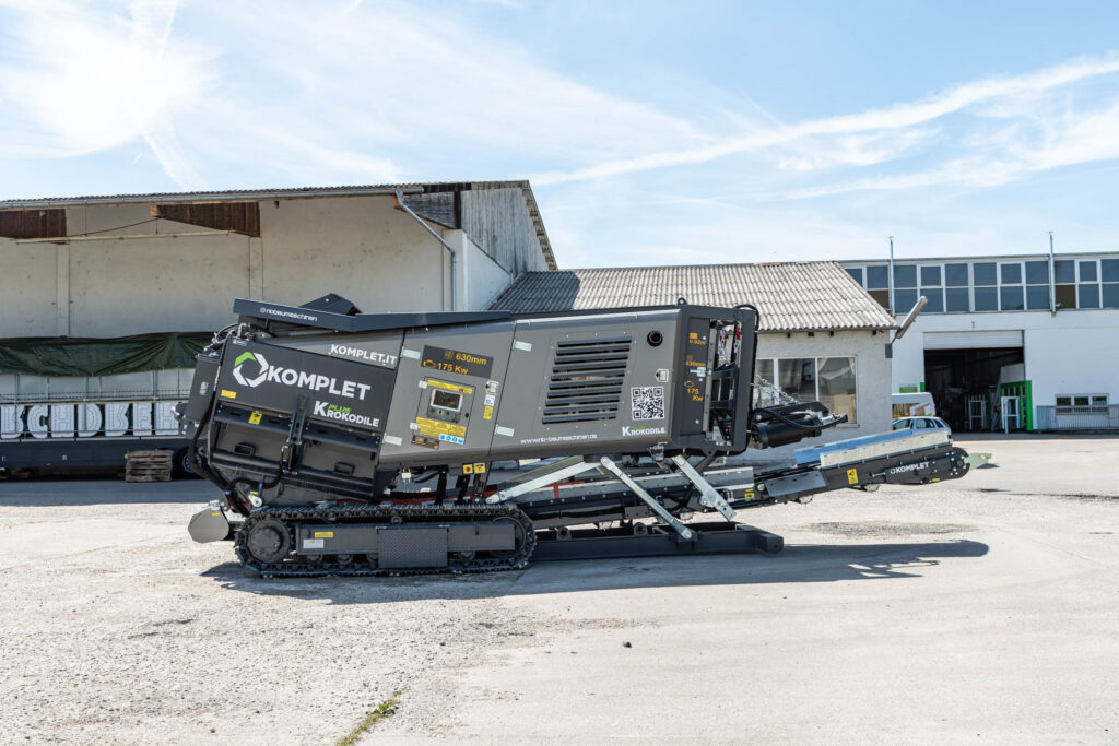 Vor Industriebauten steht ein grauer Komplet Krokodile PLUS Schredder auf Raupenfahrwerk, mit seitlichen Lüftungsgittern und ausfahrbarem Förderband.