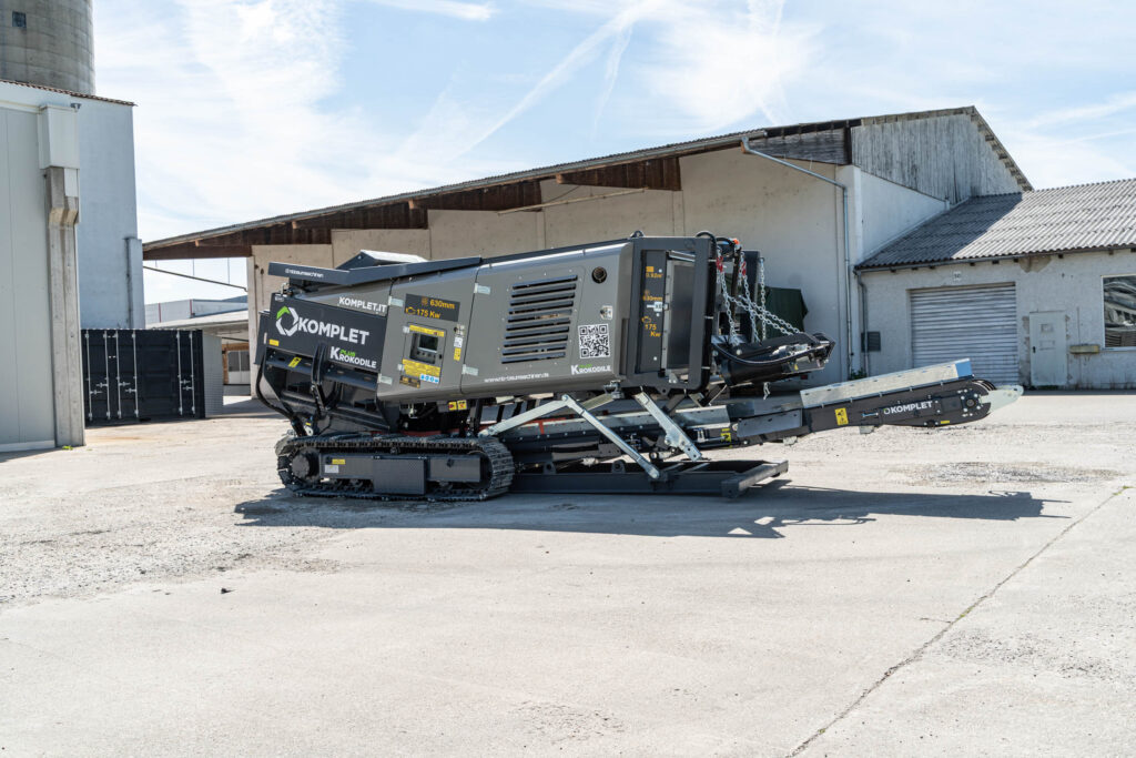 Grauer KOMPLET Krokodile PLUS Schredder auf Raupenfahrwerk steht auf einem Hof vor Industriegebäuden, mit ausgefahrenem Förderband und Ketten am Gerät.