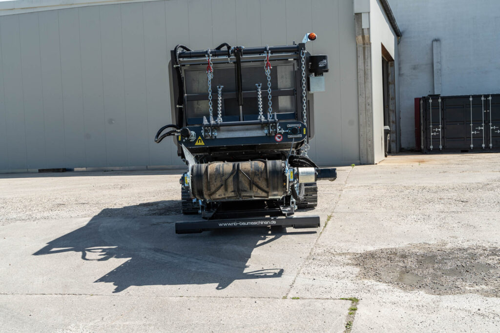Ein schwarzer mobiler Schredder steht vor einer hellen Industriehalle, mit Ketten, Raupenfahrwerk und großer Trommel im Freien.